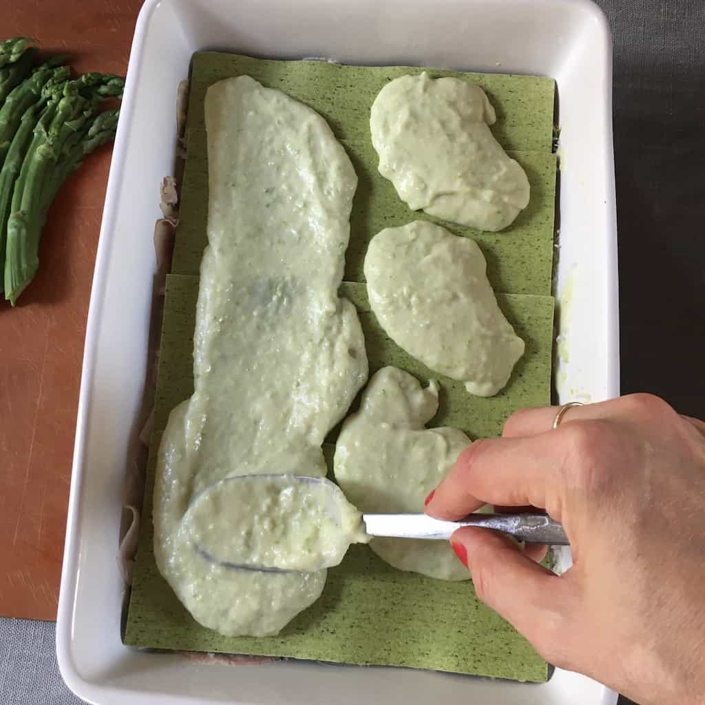 my hand using a spoon to smooth out the green bechamel over the tops of the green pasta sheets inside of a white ceramic lasagna that's lying on top of a tan colored Epicurean brand cutting board with fresh asparagus off to the left in the background