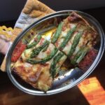 a hand holding an oval stainless steel serving platter with two orange and white plaid pot holders underneath and filled with two very generous servings of golden lasagna with bright green asparagus spears decorating the top and a red pork sauce peeking out from underneath the lasagna