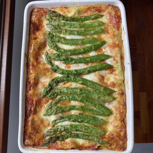 a freshly baked, crispy gooey cheesy lasagna with beautifully placed spears of asparagus on top curving out from the center going left and right
