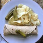 halved tuna fish salad wrap on a plate with plain potato chips, a pickle and pickled okra