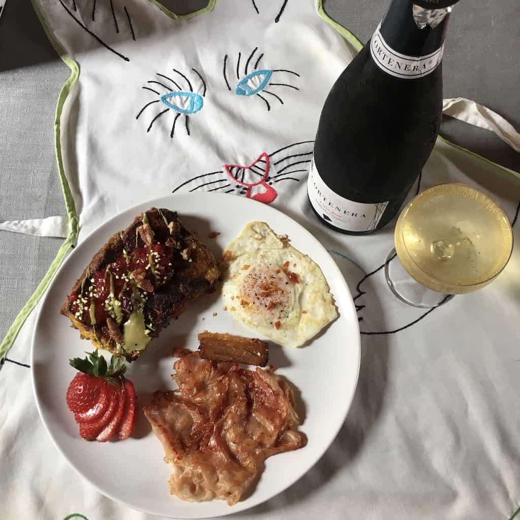 a crazy bunny apron with eyes very close together laid out on a table with grey table cloth and a plate with ham, eggs, strawberries and french toast next to a bottle of Cortenera bubbly and a vintage champagne glass filled to the top