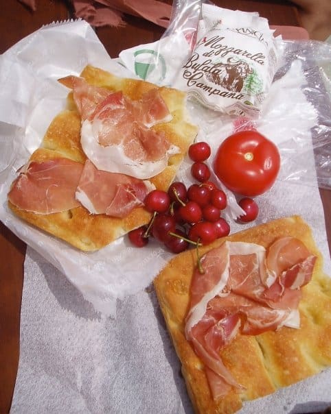 two slices of focaccia with each a slice of prociutto di Parma draped on top, a fresh ball of mozzarella di bufula still in the white package lying above with a handful of fresh dark cherries in the middle and a ripe tomato laid out on thin butcher-like paper outside for a picnic on a very sunny day