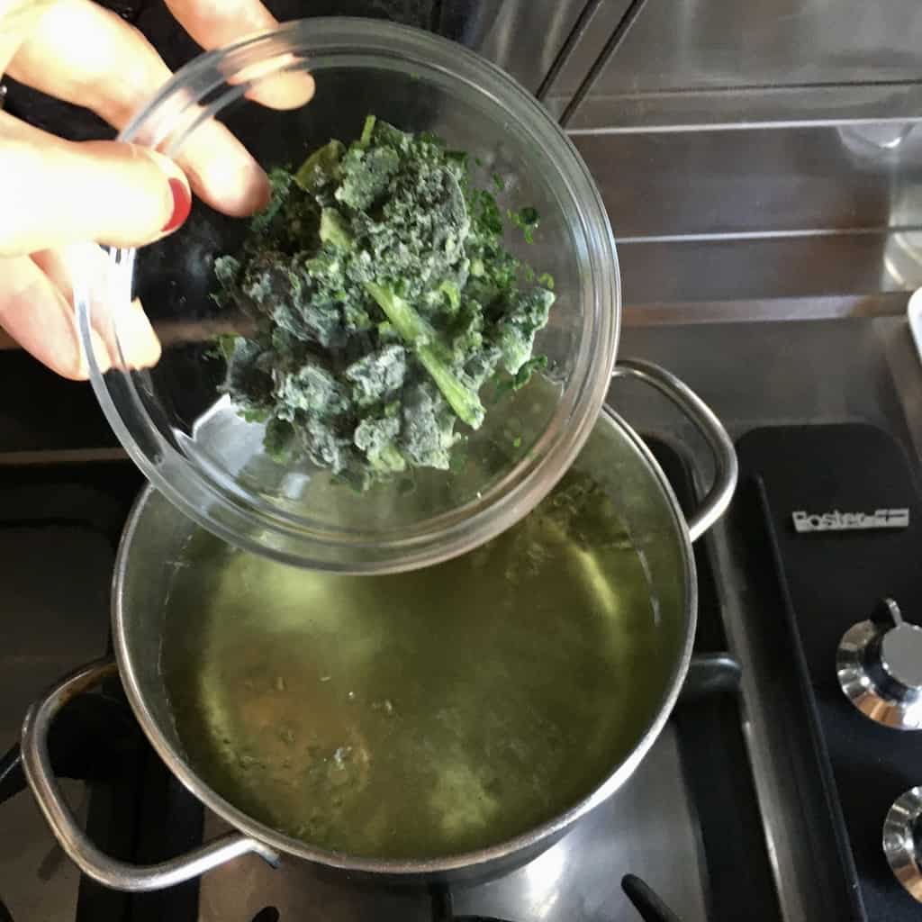 placing in the frozen spinach leaves to blanch them (hand holding the clear glass bowl with the spinach about to turn them out into the pot of slightly asparagus-stained green boiling water