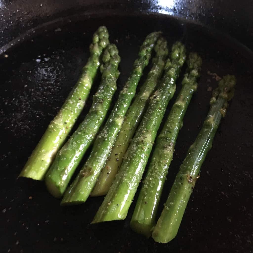 asparagus spears cooking in cast iron black pan all lined up next to each other in a row and glistening with olive oil, salt and black pepper