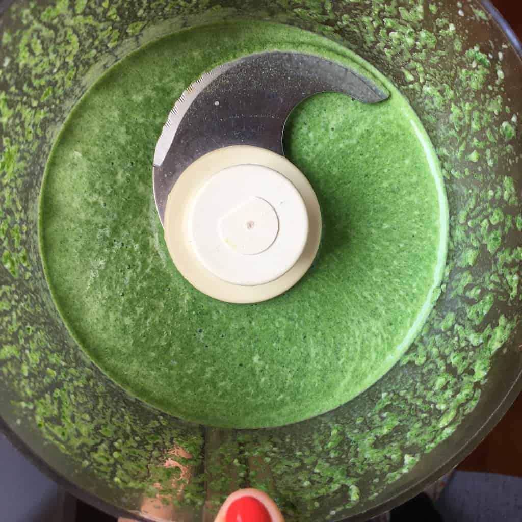 top down view of very bright green mixture in food processor bowl after being processed with tip of red painted thumbnail holding the bowl