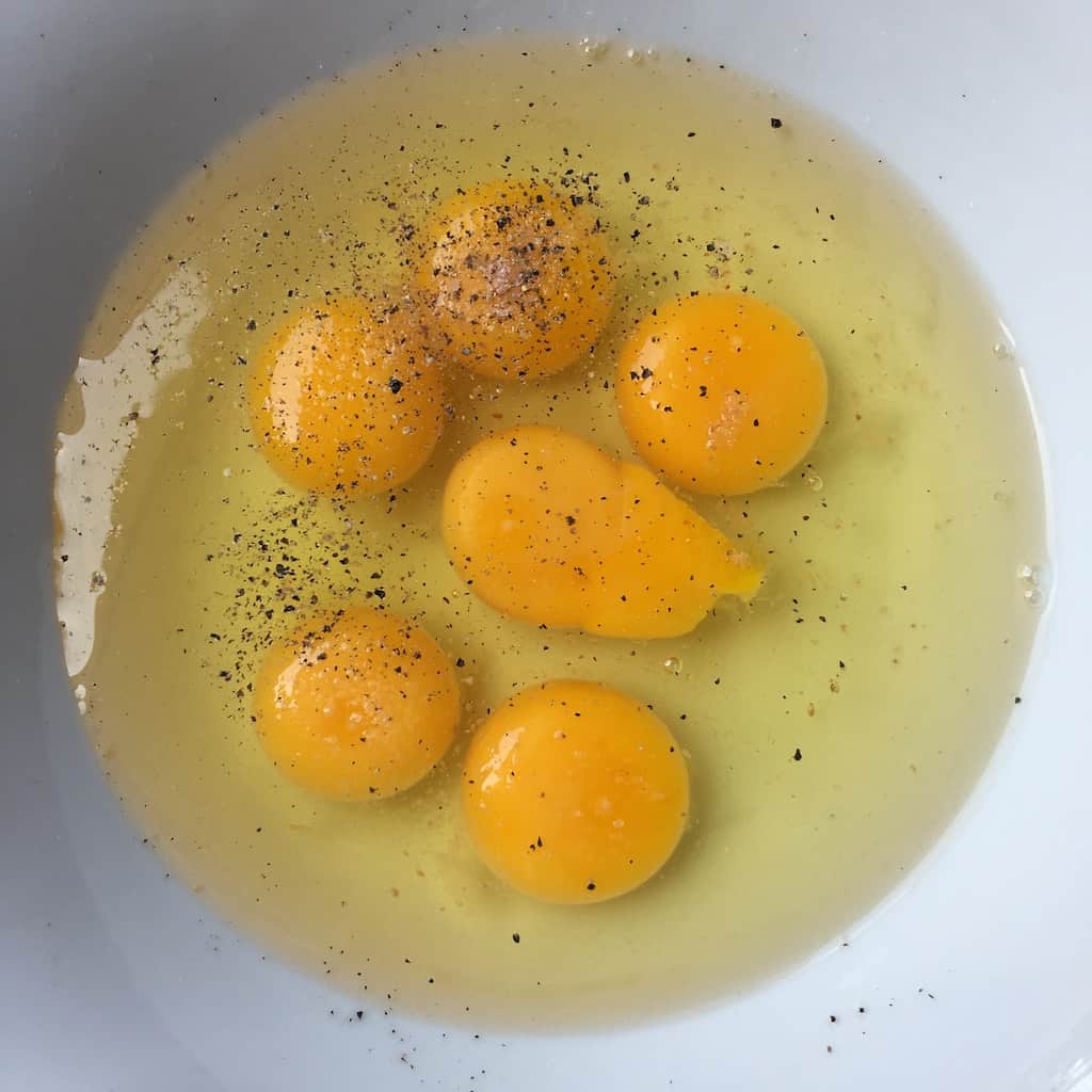 6 eggs cracked into a white bowl with salt and freshly cracked black pepper