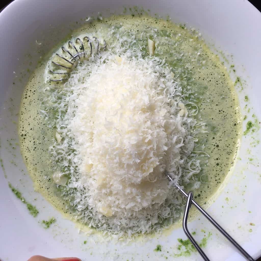 all of the green mixture poured into the 6 eggs cracked into a white bowl with salt and freshly cracked black pepper after being whisked with a pile of grated cheese added on top