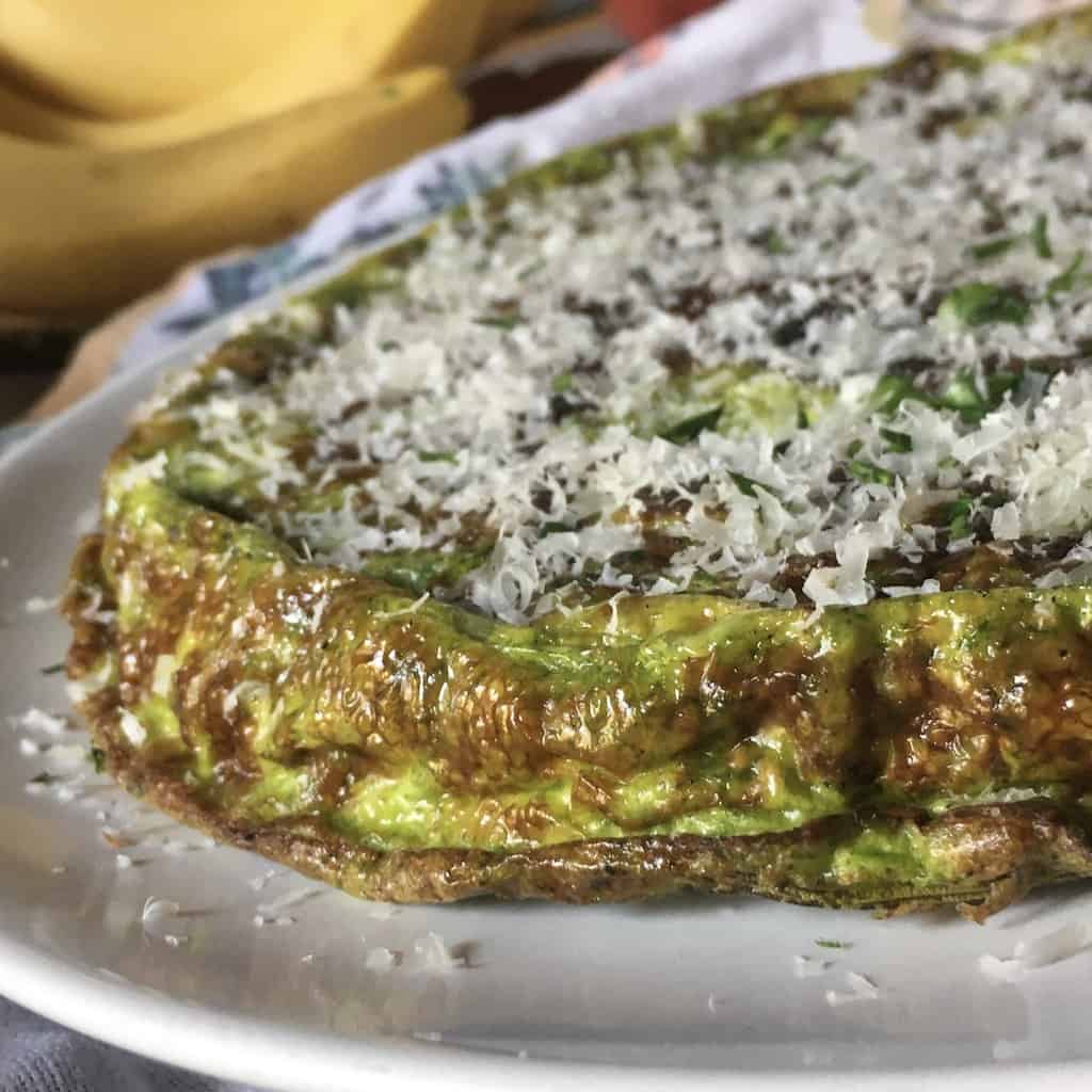 Beautifully golden brown green hued spring asparagus and zucchini frittata glistening with olive oil and dusted with freshly grated Grana Padano cheese on a white serving plate perfect for Easter brunch, lunch or easter dinner. 