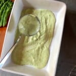 Pastel colored green asparagus bechamel sauce being spooned into a lasagna dish showing it's creamy texture and bright spring color for layered Mother's Day lasagna.