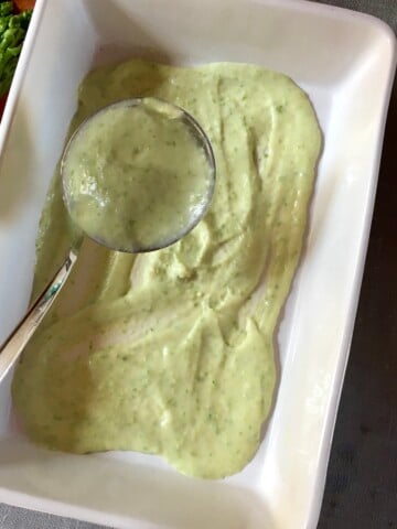 Pastel colored green asparagus bechamel sauce being spooned into a lasagna dish showing it's creamy texture and bright spring color for layered Mother's Day lasagna.