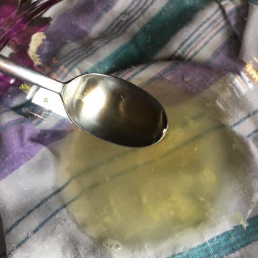 a glass bowl with lemon juice in it and a spoon above it to catch any seeds all resting on top of a linen tea towel with turquoise and lilac striped pattern