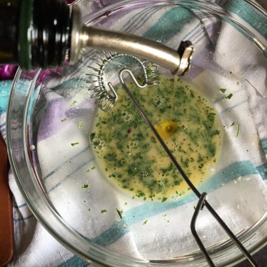 whisking the lemon and herb mixture with a flat coil whisk while drizzling a bottle of olive oil into the bowl