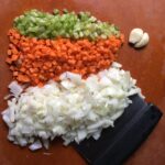 piles of mirepoix or diced onion, carrot, celery and two peeled and smashed garlic cloves on a cutting board with a dough scraper under a portion of the onions