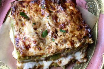 a closeup of an oozing hot square of lasagna Bolognese slice with layers of spinach egg pasta, ragù alla bolognese sauce, bechamel and Parmigiana cheese on a piece of brown parchment paper on top of a gold and pink tray