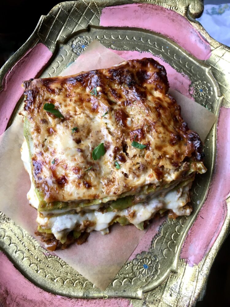 a closeup of an oozing hot square of lasagna Bolognese slice with layers of spinach egg pasta, ragù alla bolognese sauce, bechamel and Parmigiana cheese on a piece of brown parchment paper on top of a gold and pink tray