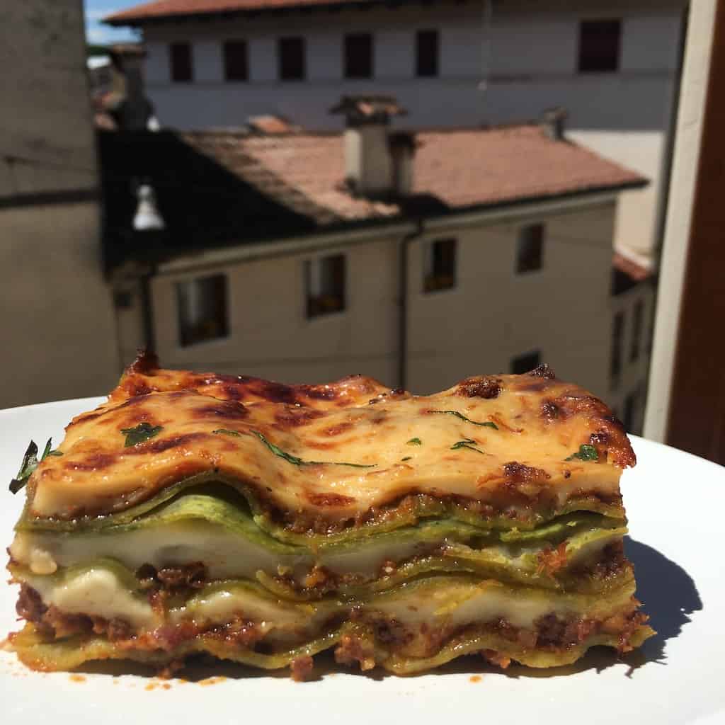 a portion of lasagna bolognese on a white plate with a view of Italian homes in the background and blue skies for days