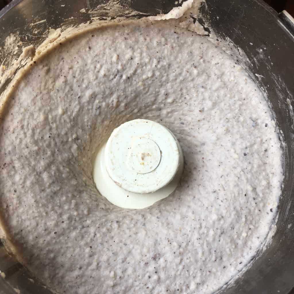 a view looking down into the food processor of the blackeyed pea hummus freshly processed into a smooth, creamy grey-purple-pink'ish hued mixture with slight texture of toasted sesame seeds