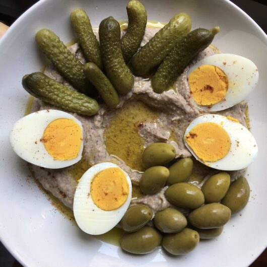 black-eyed pea hummus in the middle of white platter with a pool of extra virgin olive oil in the middle with two halves of a hardboiled egg on one side of the outer rim of hummus and another two halves opposite and on one other side a handful of green olives with pits from Puglia, Italy and on the other side a handful of mini cornichons ready to be eaten