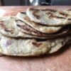 a stack of whole wheat "Everything" naan bread with blisters all over the pieces from the heat of the cast-iron skillet while cooking resting on an Epicurean Brand brownish-tan cutting board