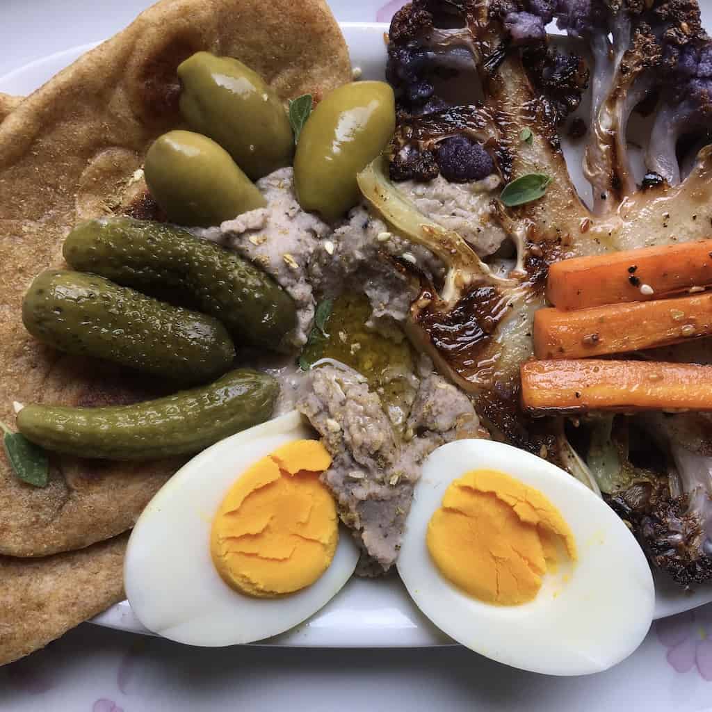 mezze platter with black-eyed pea hummus filled with olive oil, a hardboiled egg halved, sauteed caramelized carrot sticks, pan-seared purple cauliflower "steak" green olives from Puglia, baby cornichons and fluffy whole wheat naan bread on a white platter