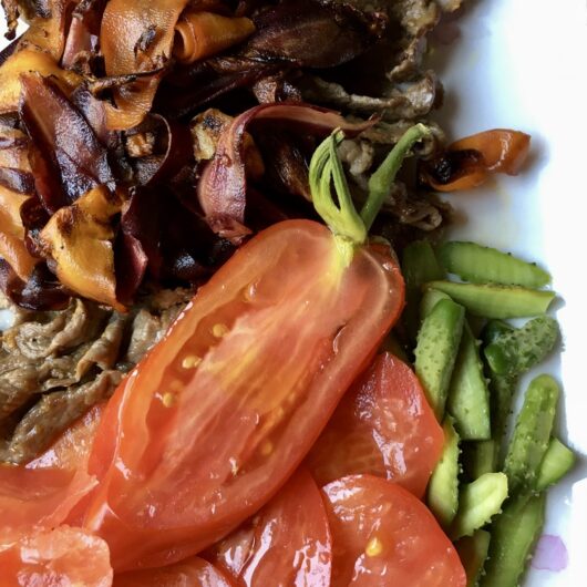 closeup of a white platter with shaved garlic ginger beef and caramelized purple and orange carrot ribbons on top with thin vertical slices of long cornabel tomatoes and the smallest baby fresh cucumbers you've ever seen also sliced vertically like the tomatoes
