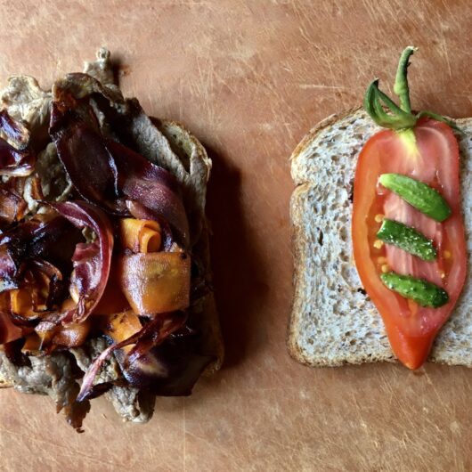 two halves of a garlic-ginger shaved beef sandwich lying on a cutting board - one half of made with whole wheat bread slices is covered in shaved beef, caramelized purple carrots and orange carrot ribbons and the other half has a single beautiful slice of cornabel tomato with stem still intact and 3 tiny baby cornichon style cucumbers sliced lengthwise fresh from the garden lying on the tomato from top to bottom with equal distance between them