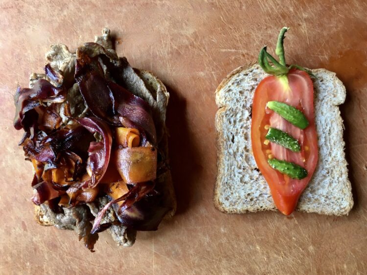 two halves of a garlic-ginger shaved beef sandwich lying on a cutting board - one half of made with whole wheat bread slices is covered in shaved beef, caramelized purple carrots and orange carrot ribbons and the other half has a single beautiful slice of cornabel tomato with stem still intact and 3 tiny baby cornichon style cucumbers sliced lengthwise fresh from the garden lying on the tomato from top to bottom with equal distance between them