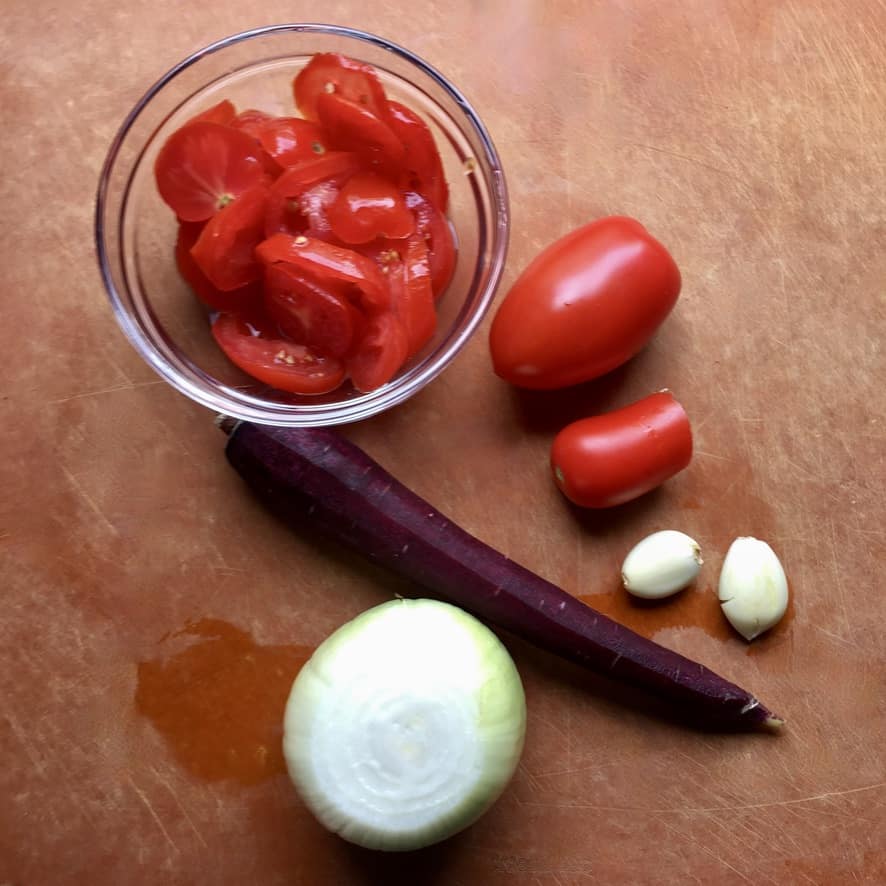 sliced tomatoes in a small glass prep bowl, a purple carrot with the skin removed, 2 peeled garlic cloves, 2 whole san marzano tomatoes, and 1 whole white onion with the peel removed all resting on a cutting board