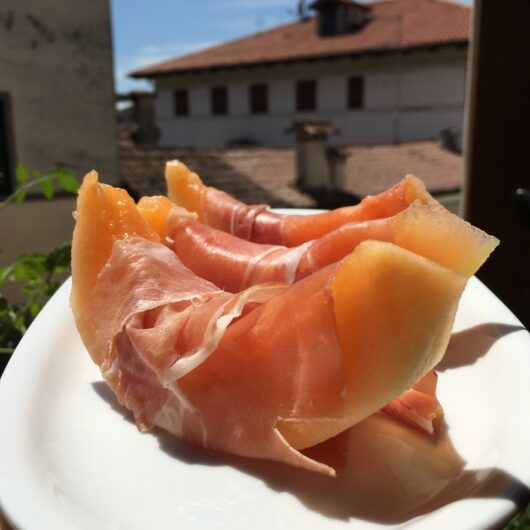 cantaloupe wedges draped and wrapped with beautifully soft and luscious paper-thin sheets of prosciutto di parma on a marble round platter in the sunlight with a view of Italy in the background