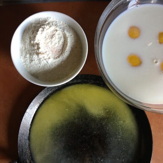 raw ingredients for crêpe batter with flour in a small white bowl, eggs floating in milk, and melted butter in the crêpe pan