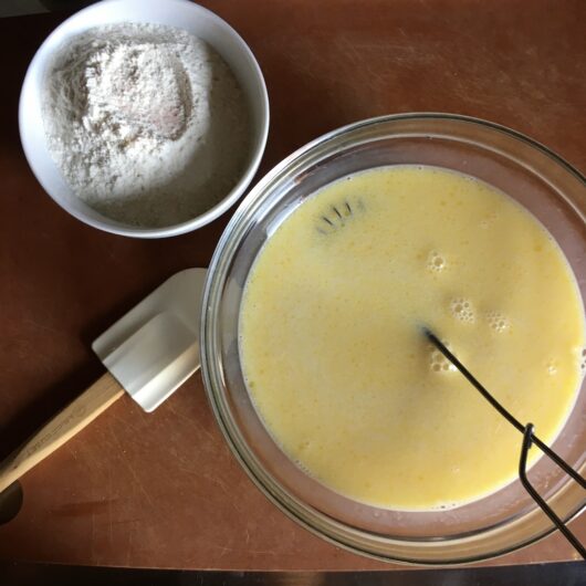 a bowl of flour with sugar and salt next to a white Le Creuset rubber spatula and a bowl of whisked wet crepe batter ingredients