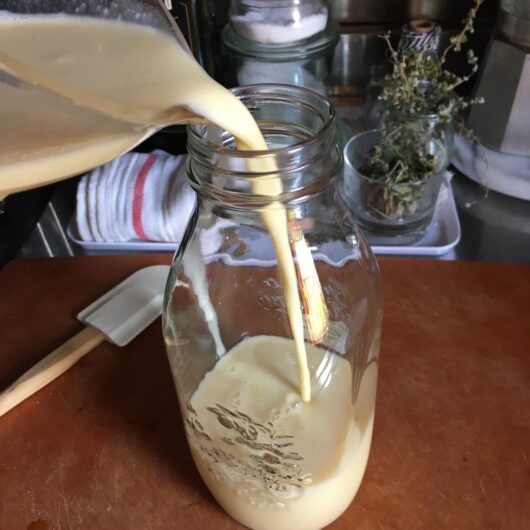pouring the crepe batter ingredients into a Quattro Stagione 1 liter tall glass canning jar with a spatula lying next to the bottle
