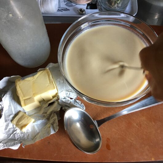 the next day the crepe batter poured into a medium sized glass mixing bowl and being whisked with and opened package of butter to the left of the bowl, and a ladle lying in front of the mixing bowl with the empty bottle in the background