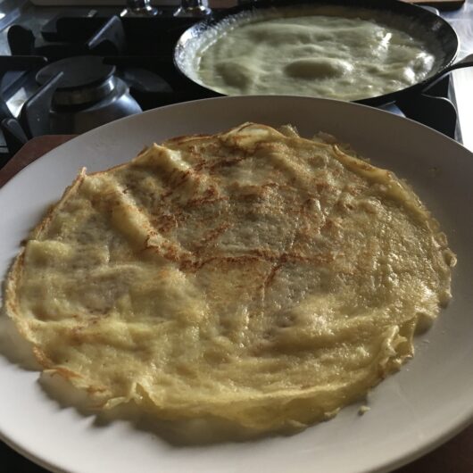 side shot of a white plate with freshly cooked crepes on it and a view of the crepe pan cooking another crepe