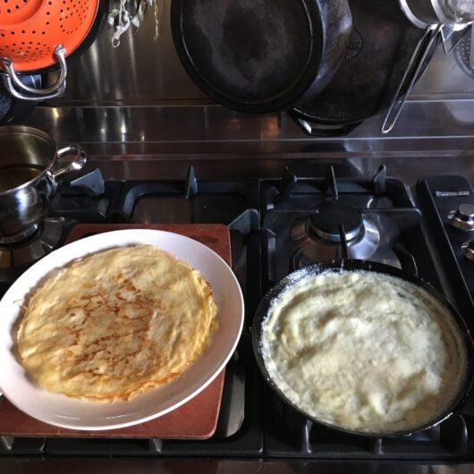 top down shot of a white plate to the left of freshly cooked crepes on it and a view of the crepe pan cooking another crepe