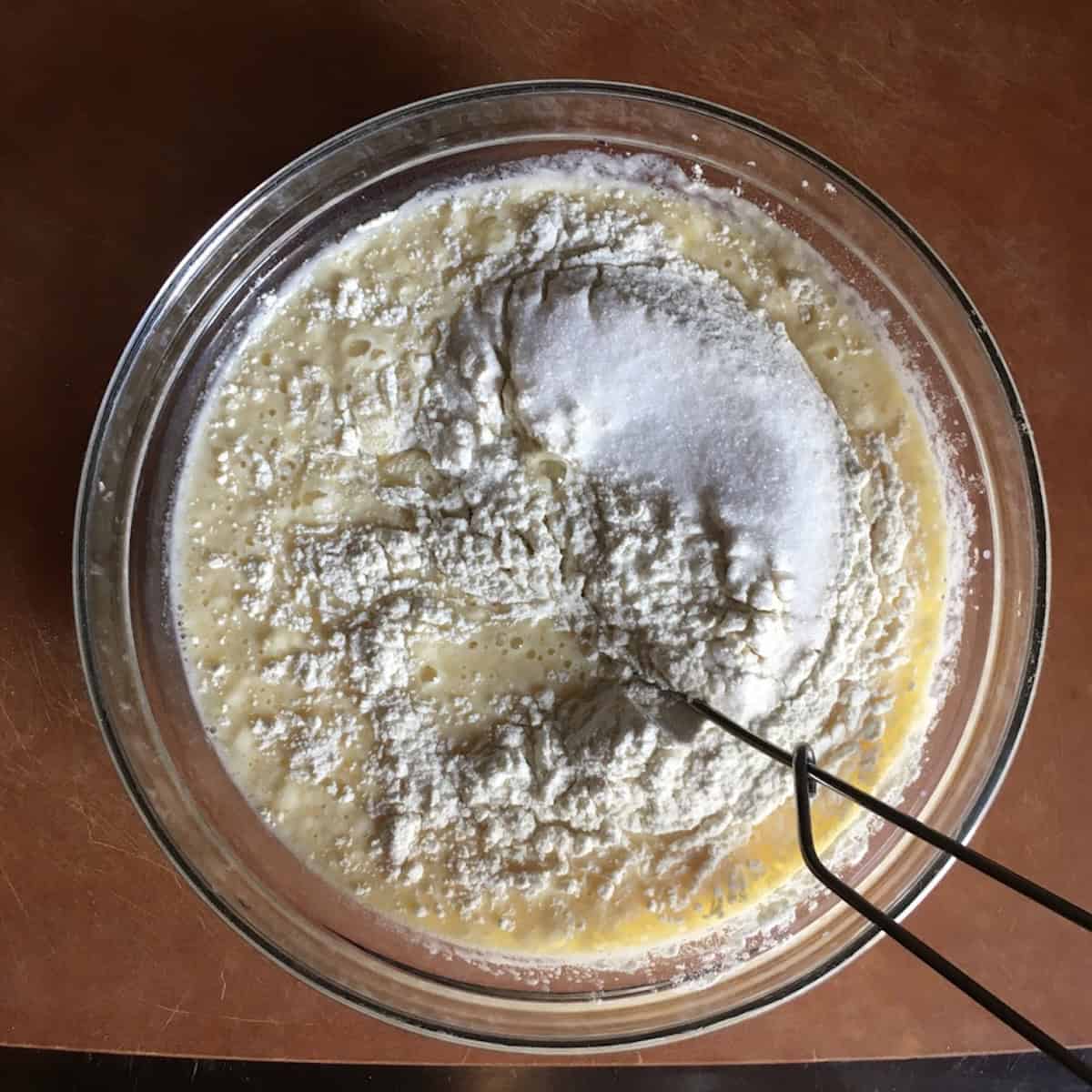 Flour and sugar mixture added to eggs, milk, and browned butter in a mixing bowl before whisking, a key crepes ingredients step for this simple crepe batter.
