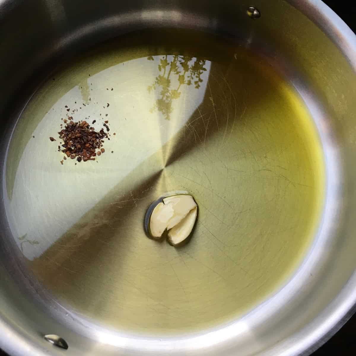 Garlic clove and crushed red pepper in a stainless steel saute pan ready to blister for clam sauce recipe.
