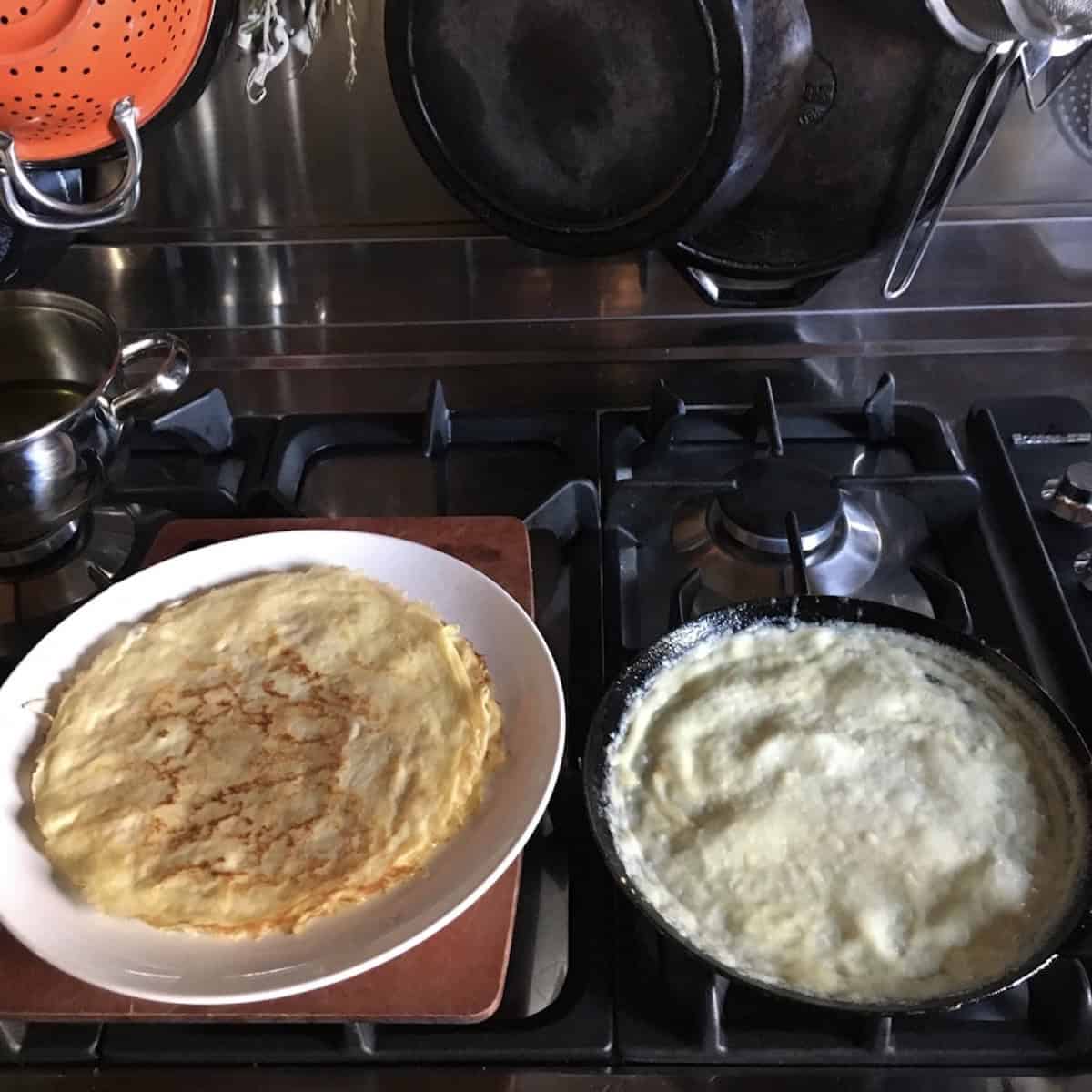 A stack of cooked crepes on a plate beside a crepe pan with one crepe still cooking, showing how to cook crepes and how long to cook crepes for this basic crepe recipe.
