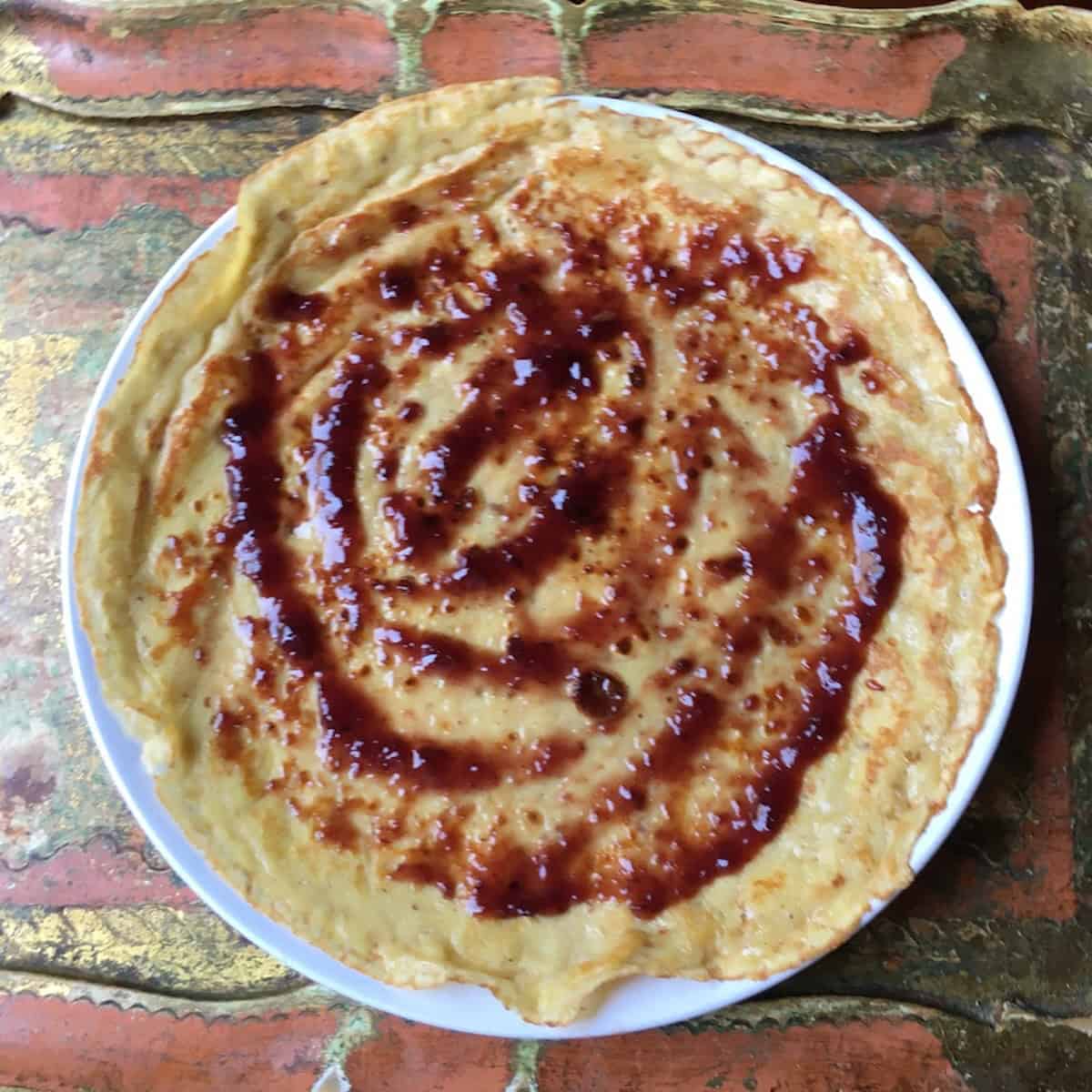 Cherry jam spread across a crepe with an offset spatula, a simple sweet crepe recipe and crepe filling ideas for this crepe filling recipe.
