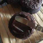 a dollop of shiny dark chocolate ganache in the middle of a chocolate cupcake with a small cake spatula spreading the ganache evenly over the top of the cupcake