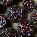 closeup of a tray full of devil's food cake cupcake next to the others frosted with a thin layer of glistening dark chocolate ganache frosting with pink, turquoise, lilac and orange jimmies and green balls with a few nonpareil sprinkles