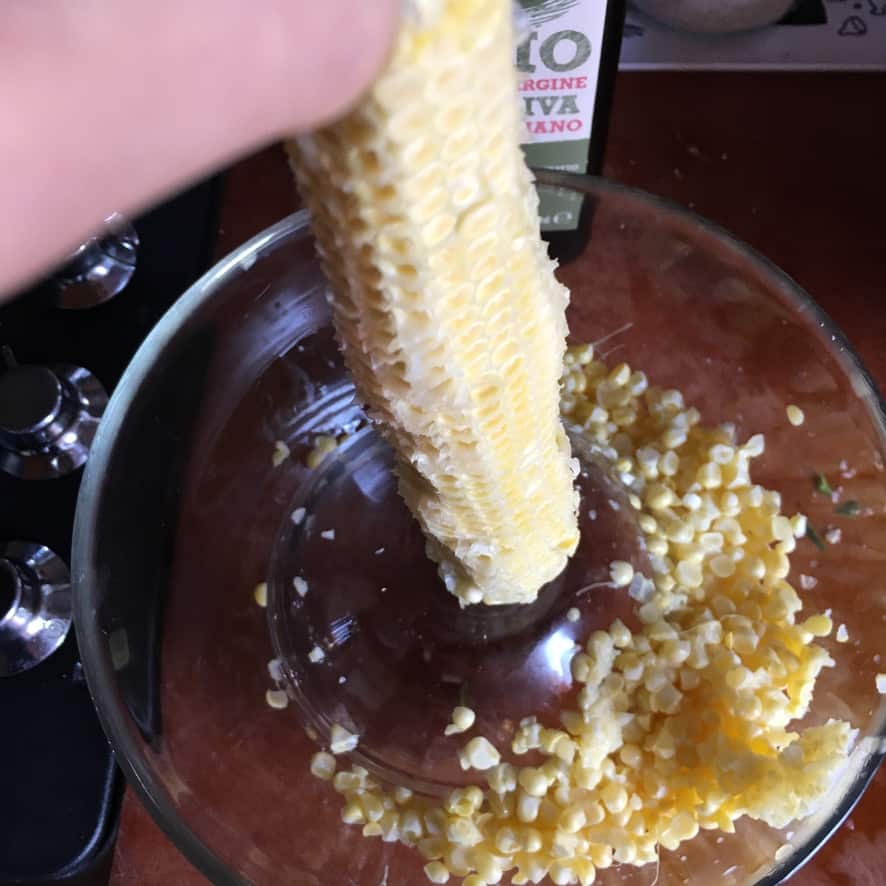 a glass mixing bowl with a small glass mixing bowl turned upside down inside the bowl with an ear of corn resting on the small upside down bowl to allow the kernels to fall into the bigger bowl as they are sliced off the cobb