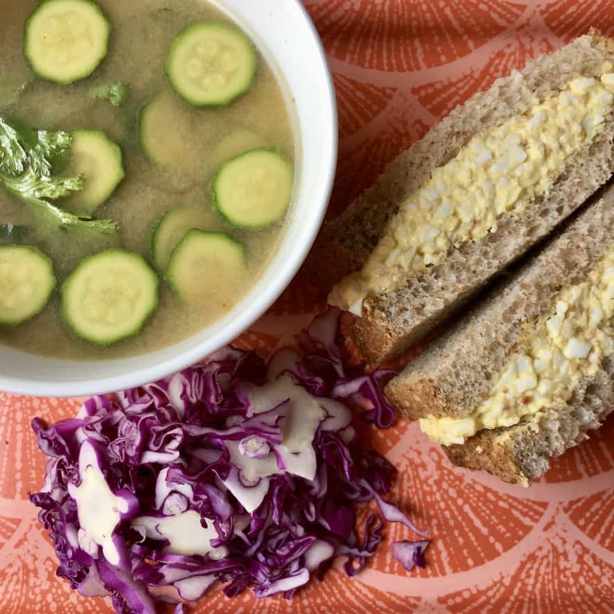 halved egg salad sandwich on a coral Japanese inspired tray with a bowl of miso soup with zucchini rounds floating at the top and a small salad of purple cabbage next to it