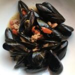 steamed black Sardinian mussels with bits of sausage and mushrooms and diced tomatoes in a white wine broth in a white bowl (top down view)