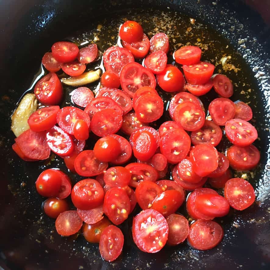 raw sliced baby grape Datterino tomatoes in olive oil with a smashed garlic clove in a black pan