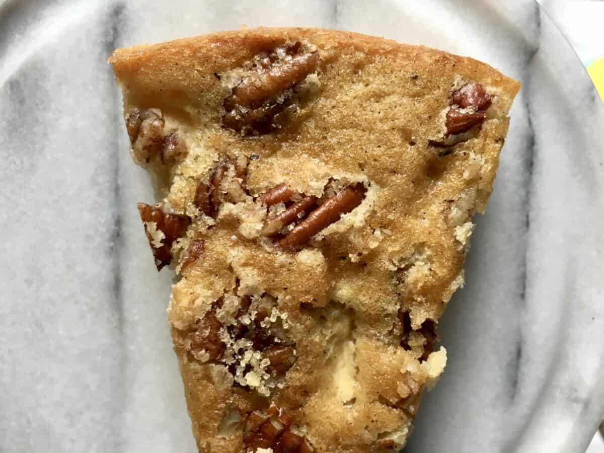 a slice of Apple rum cake with a golden crunchy pecan topping next to another slice on a sheet of white parchment paper in the sunlight