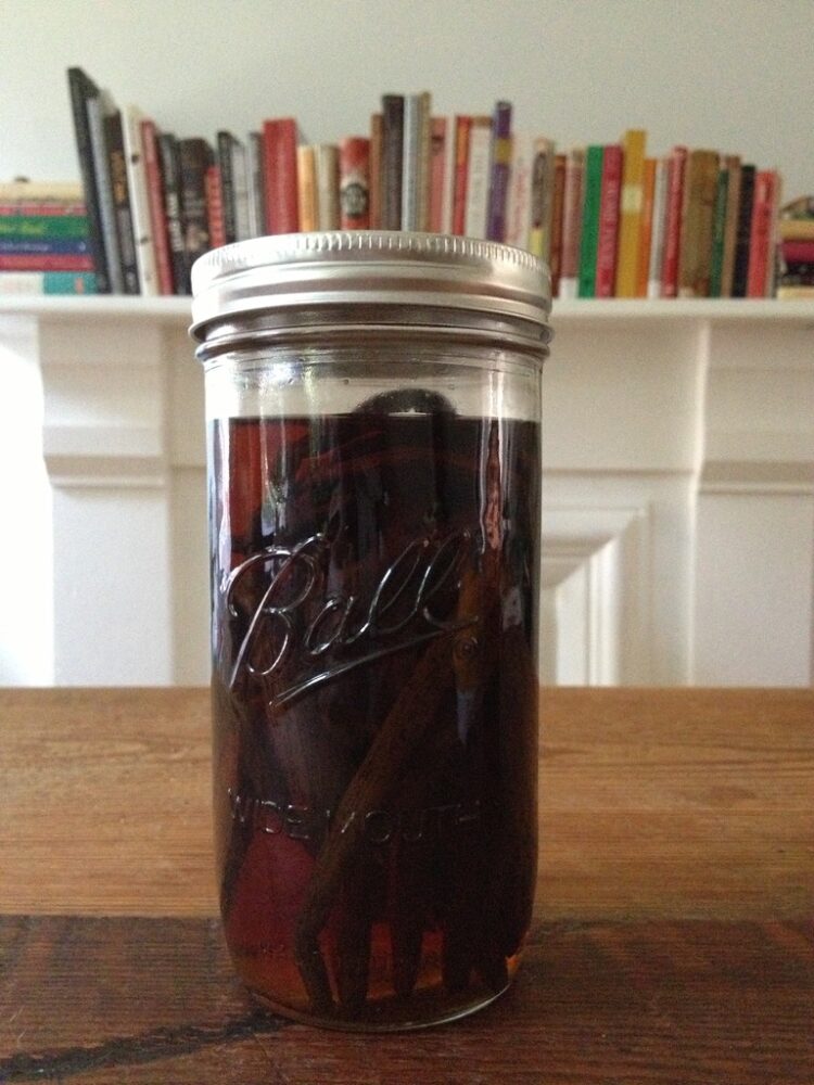 a tall wide mouth Ball Mason Jar with silver lid and band full of dark vanilla beans covered in vodka and looking dark and beautiful like an extract on top of a wooden dining table with the white marble fireplace lined with a row of cooking books from end to end in my old brownstone in Brooklyn