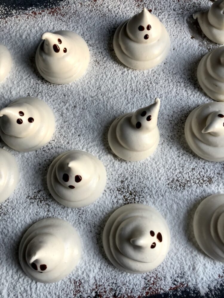 a sheet tray dusted with powdered sugar and ghost-shaped marshmallows piped onto it with dark chocolate eyes and mouths