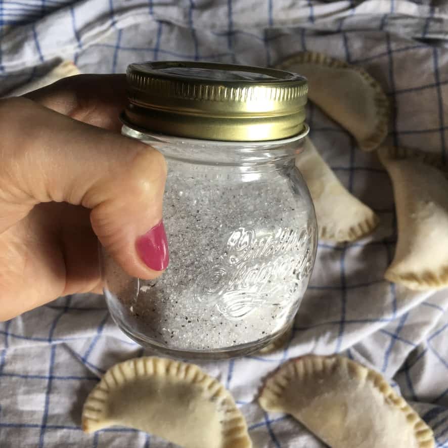 me holding an Italian jam glass canning jar with gold lid holding vanilla bean speckled white sugar with raw fried pies in the background on a blue and white checkered linen tea towel
