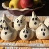 happy and angry-faced ghost meringues on an orange and white checkered tea towel with bowl of crisp red and green apples in the background